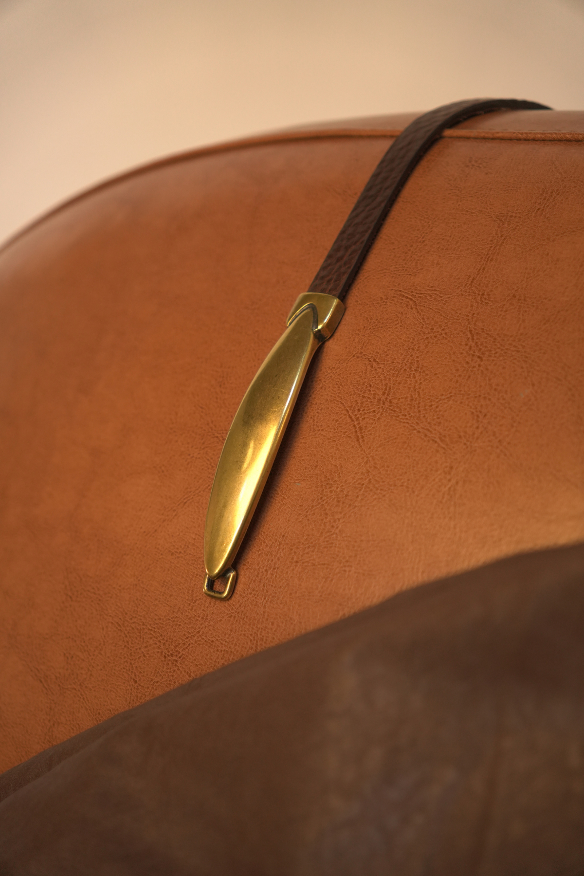 Ceinture Rachel Atelier Nappa posée sur un fauteuil en cuir : lanière marron de 1,5 cm, boucle ovale allongée et embout dorés