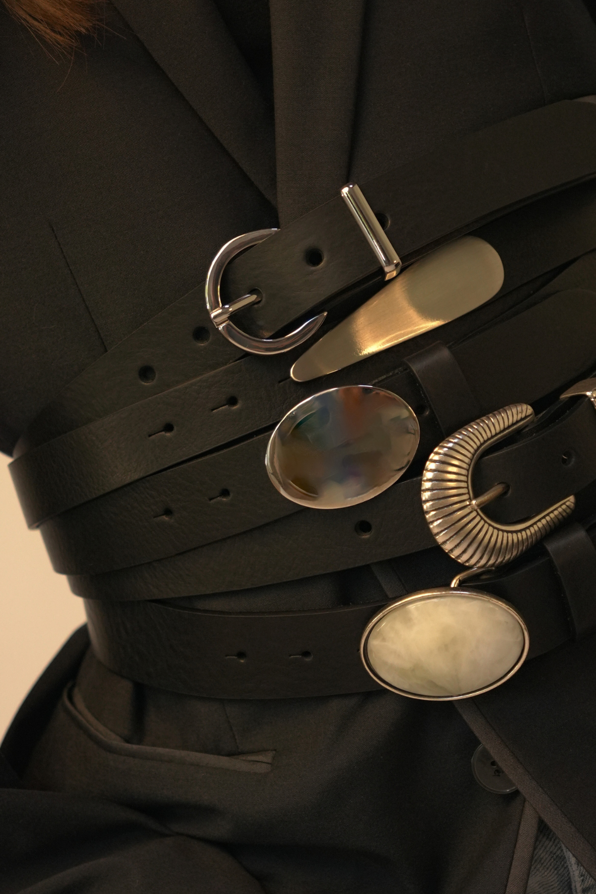 Ceintures Atelier Nappa en cuir noir superposées sur un blazer, dont la Kamila à grande boucle ovale argentée effet miroir