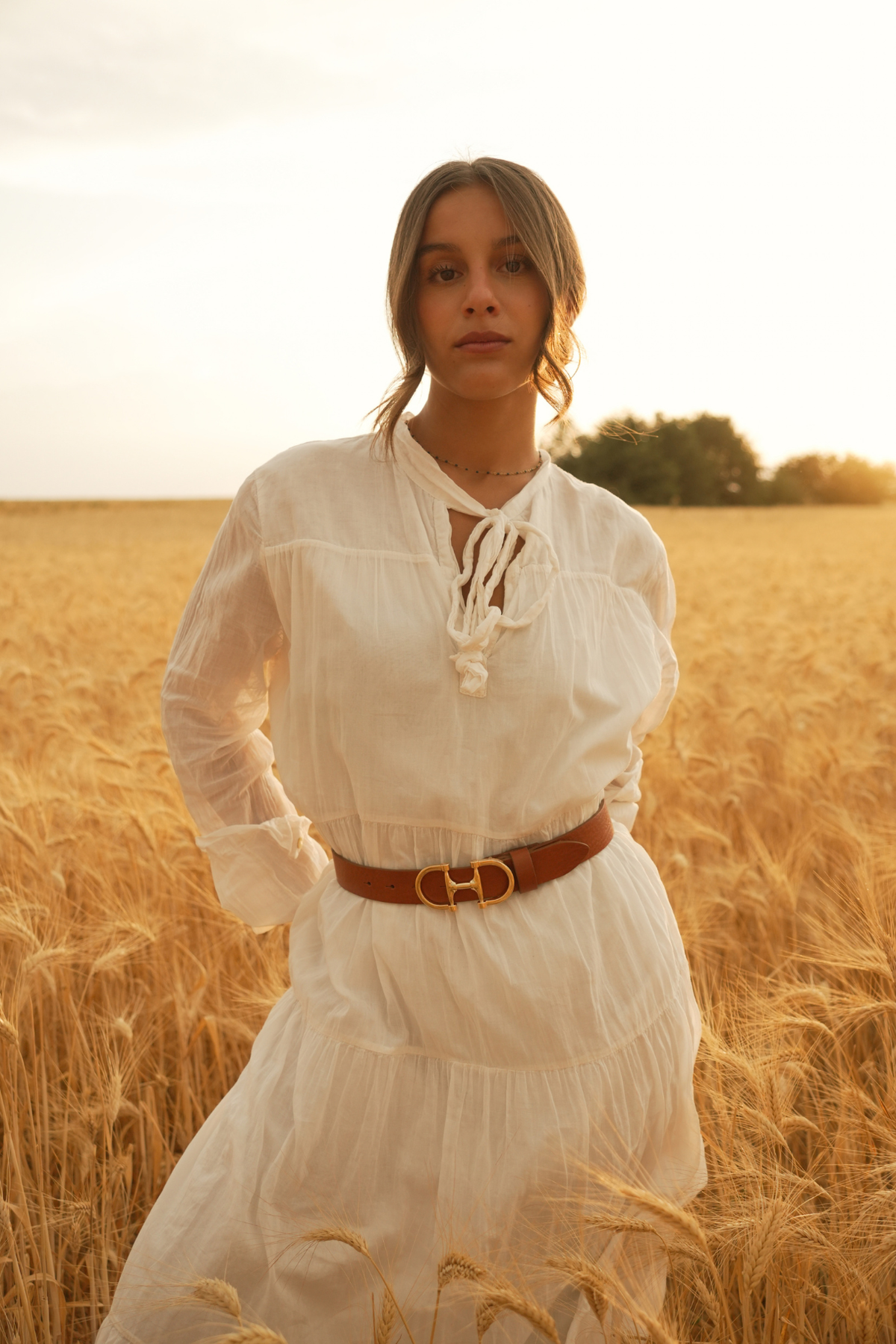 Ceinture Jeanne Atelier Nappa en cuir camel à boucle équestre dorée, portée à la taille sur une robe blanche dans un champ de blé