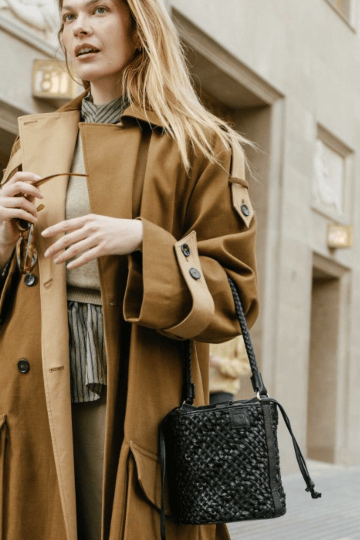 Petit sac seau en cuir tressé noir Biba Selma porté à l’épaule avec un trench